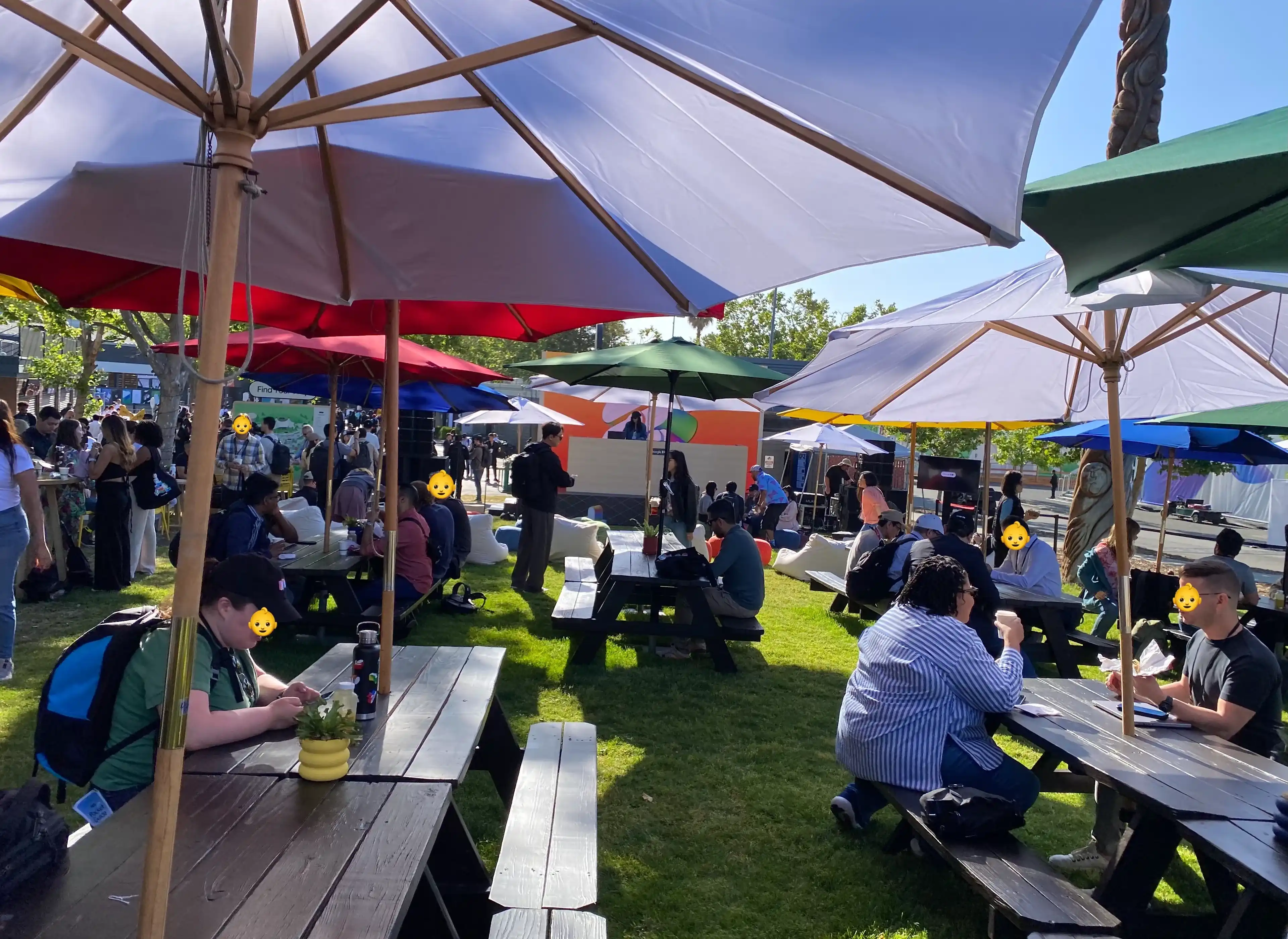 A grassy area with many benches and tables set up under parasols. There's a DJ booth with a DJ in the background, and many people are present.