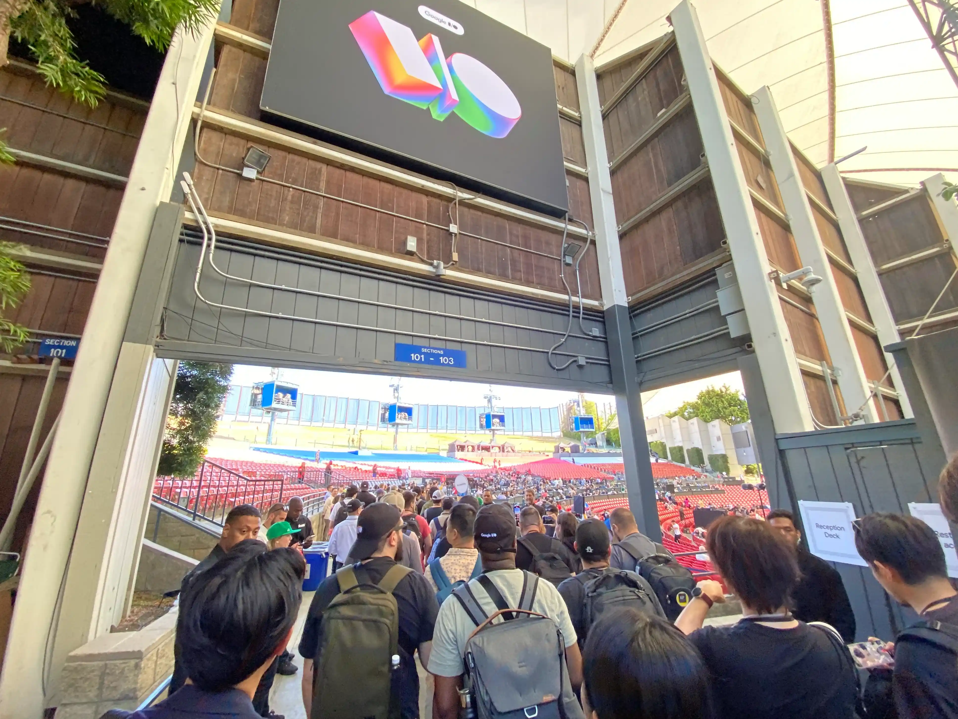 A photo of a gate-like structure with an 'I/O' sign. Beyond it is a vast venue with many seats visible. Many people are lined up, just about to enter.