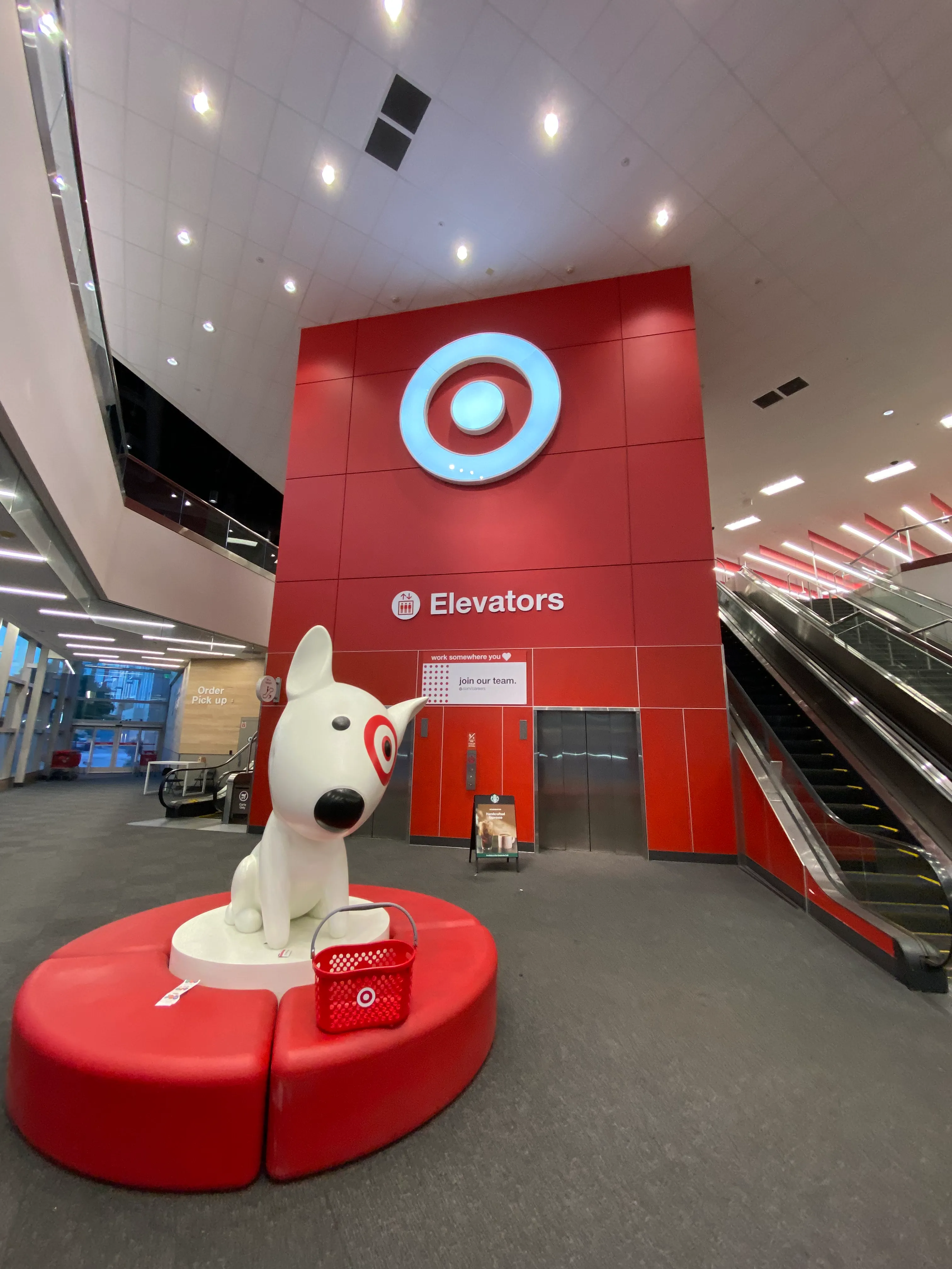 Inside a building, in a spacious hall-like area, there are two or three escalators and elevators. A large statue of Target's dog mascot is visible. No people are in the shot.