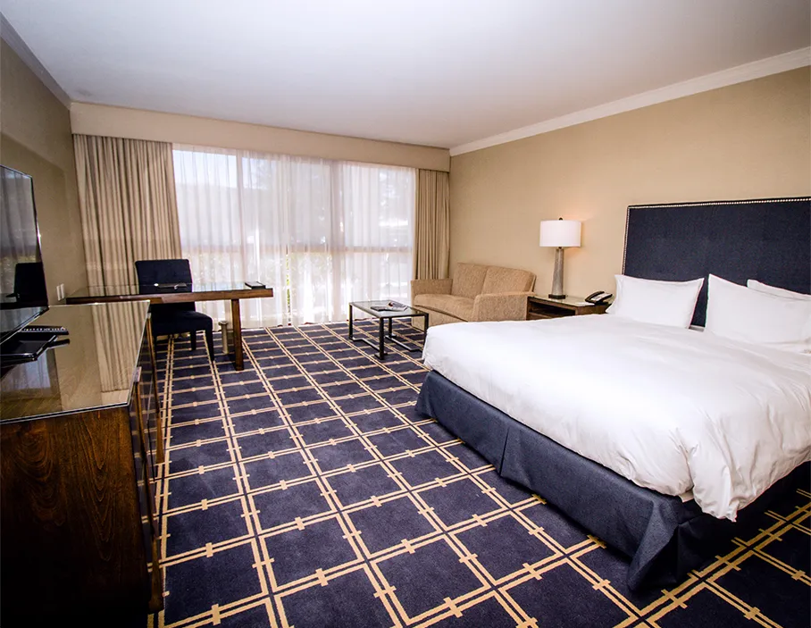 A hotel room featuring a TV and TV stand in the front left, a desk with a lamp in the rear left, and a sofa and low table in the rear right. A king bed is located in the front right, and the back wall is dominated by a large glass window.